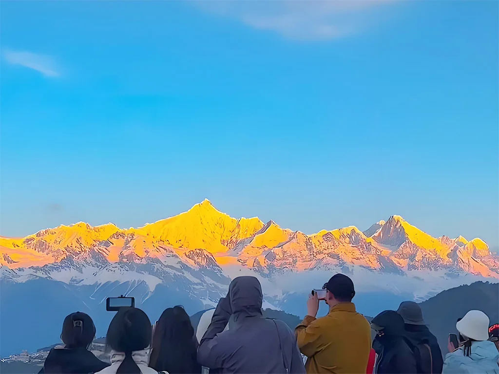 迪慶梅里雪山日照金山壯麗景觀，清晨陽光灑落在連綿雪峰上，山頂染上金色光芒，前景遊客駐足拍照欣賞壯觀山景，展現迪慶梅里雪山著名的日照金山奇景與香格里拉高原自然魅力