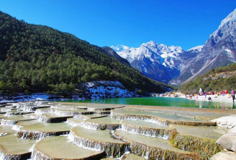 Bai Shui Terrace 玉龍雪山白水台呈現層層堆疊的白色鈣化梯田景觀,清澈流水沿著階梯狀平台緩緩流下。遠方可見雪覆的玉龍雪山與翠綠山谷,藍天映襯壯麗自然景色,遊客在湖畔與平台附近欣賞玉龍雪山白水台獨特地貌。