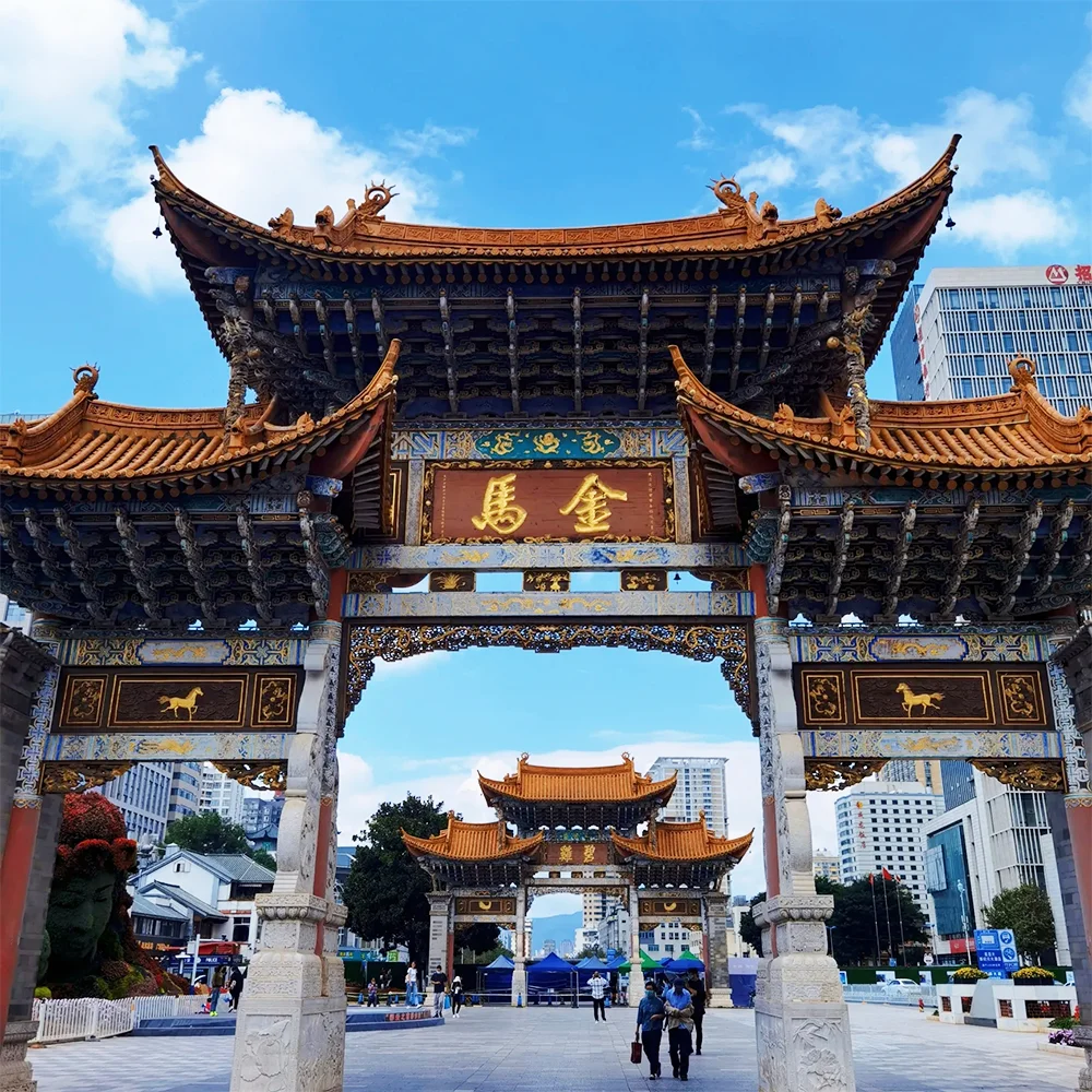 昆明金馬碧雞坊矗立於廣場，藍天下的傳統建築展現飛簷翹角與精美彩繪。牌坊刻有「金馬」字樣，與後方現代城市建築交織。