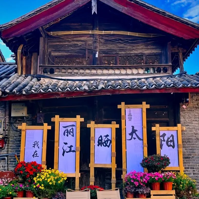 Lijiang Old Town 3 麗江古城傳統木造建築外觀,屋頂覆蓋灰色瓦片,展現納西族古城建築特色。門前擺放花卉盆栽與木製展示架,上方書寫「我在麗江晒太陽」字樣,呈現悠閒的古城生活氛圍,是遊客拍照與體驗麗江古城文化的熱門場景。