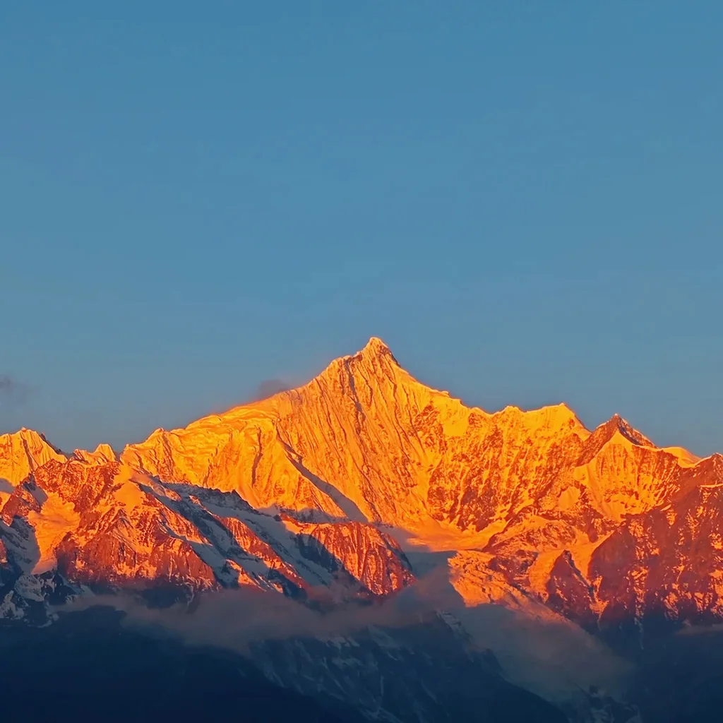 清晨陽光照耀 梅里雪山，雪峰被金色光芒染亮，山體輪廓清晰壯麗，天空湛藍，呈現震撼的「日照金山」自然奇觀。