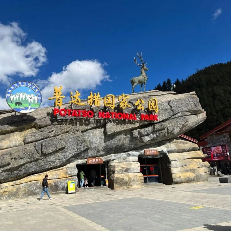 香格里拉普達措公園入口以巨型岩石造型打造，上方設有鹿雕像與「普達措國家公園」標誌字樣。藍天白雲與森林山景環繞，遊客從岩石洞口入口進入景區，呈現香格里拉普達措公園自然與生態特色的代表性地標景觀。