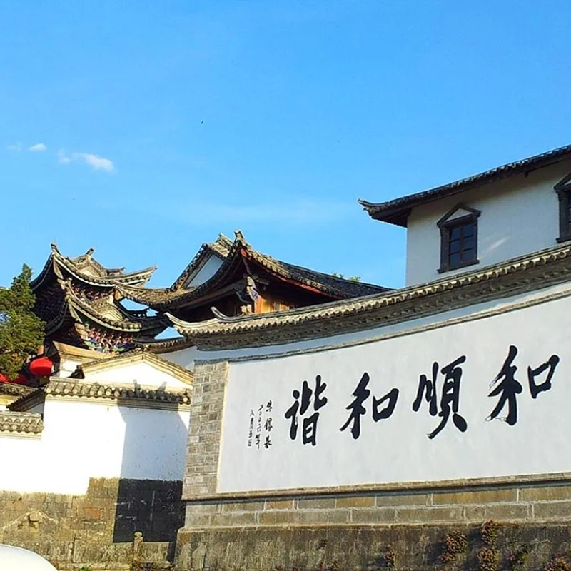 雲南騰衝的 和順古鎮 古建築景觀。白色石牆上寫著「和順和諧」書法字樣,牆後可見傳統中式屋頂與飛簷建築,青瓦層疊,展現典型的古鎮風格。晴朗藍天下,古鎮街巷寧靜雅致,整體畫面呈現濃厚的歷史文化氣息與雲南傳統人文風貌。