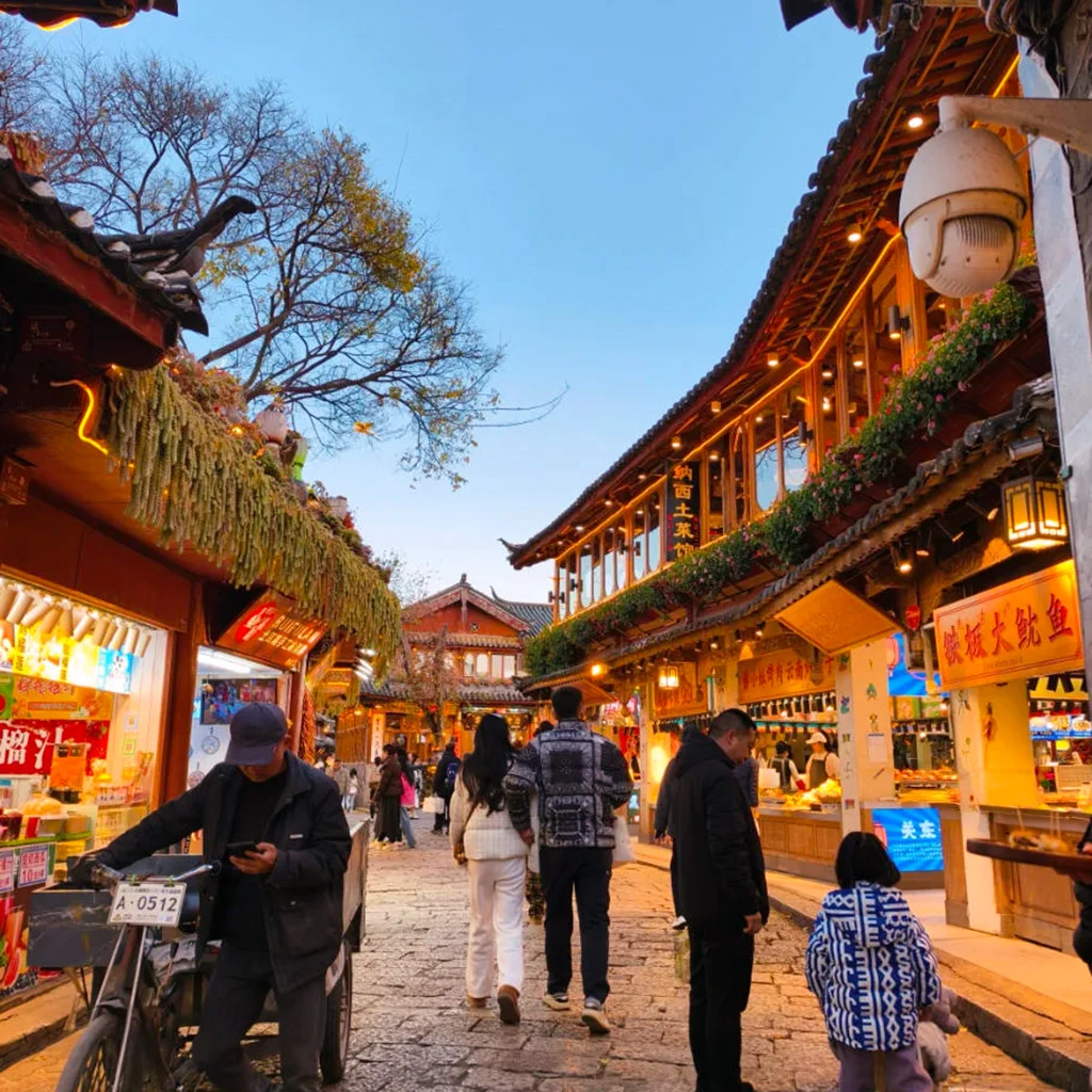 Lijiang Old Town 雲南著名歷史景區 麗江古城 的傍晚街景。石板路兩側是傳統木質建築與商店,屋簷掛著燈飾與招牌。遊客與當地居民在古街上漫步購物,街道燈光溫暖,營造出熱鬧又富有文化氣息的古鎮氛圍,展現麗江古城獨特的旅遊魅力