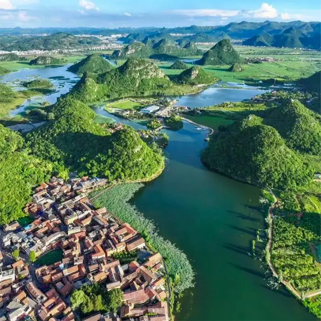 Puzhehei 航拍視角展示文山普者黑的喀斯特山水景觀。湖泊與河道蜿蜒穿過綠色山丘,湖邊分布紅瓦屋頂村落與農田,遠處群山連綿,呈現雲南典型的山水田園風光。
