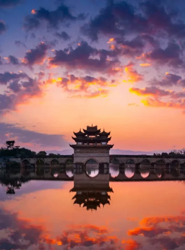 建水雙龍橋夕陽景觀，古老石拱橋橫跨水面，橋中央的傳統亭閣在晚霞映照下形成優美剪影，橋洞與天空雲彩倒映在平靜水面上，呈現雙龍橋典雅壯麗且充滿歷史韻味的古橋風光