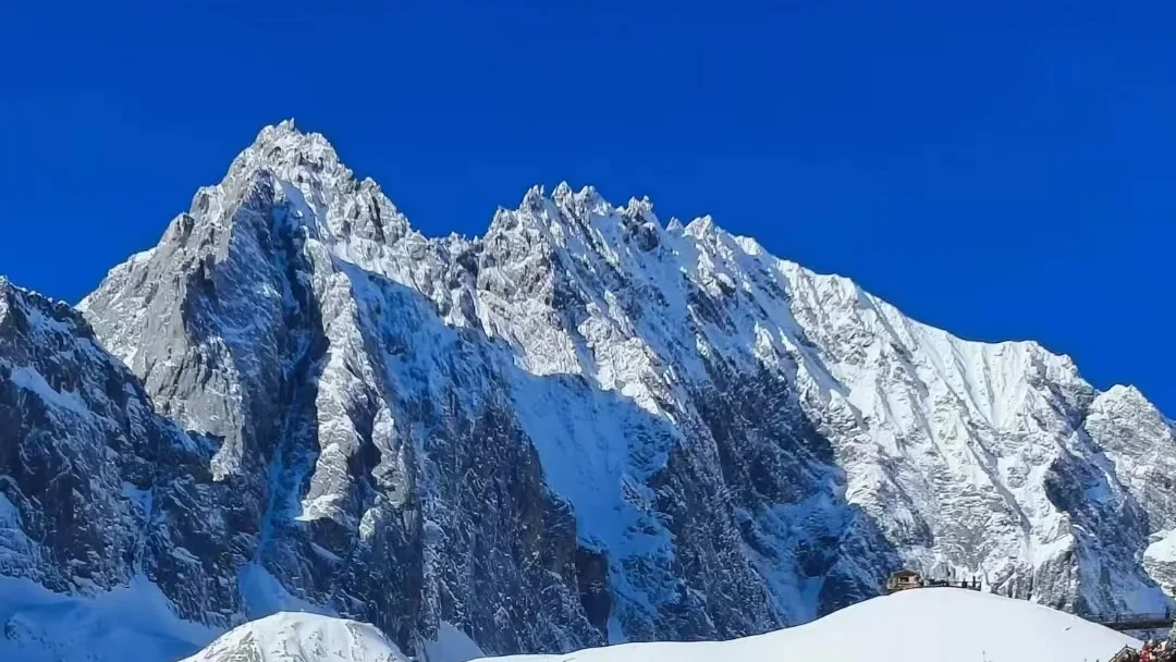雲南麗江玉龍雪山,雄偉鋸齒狀積雪山峰聳立於澄澈深藍天空下,山腳有小型觀景平台。