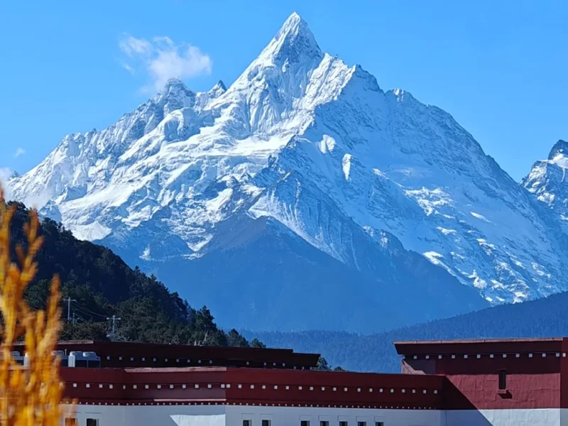 在 飛來寺 遠眺，雪山在陽光照耀下潔白耀眼，山峰巍峨聳立，前景建築與遠山形成層次分明的壯麗景觀。
