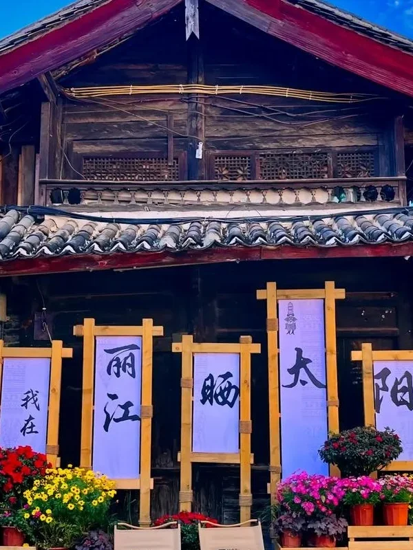 Lijiang Old Town 3 麗江古城傳統木造建築外觀,屋頂覆蓋灰色瓦片,展現納西族古城建築特色。門前擺放花卉盆栽與木製展示架,上方書寫「我在麗江晒太陽」字樣,呈現悠閒的古城生活氛圍,是遊客拍照與體驗麗江古城文化的熱門場景。