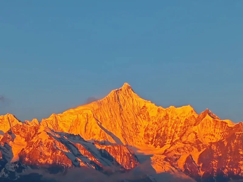 清晨陽光照耀 梅里雪山，雪峰被金色光芒染亮，山體輪廓清晰壯麗，天空湛藍，呈現震撼的「日照金山」自然奇觀。