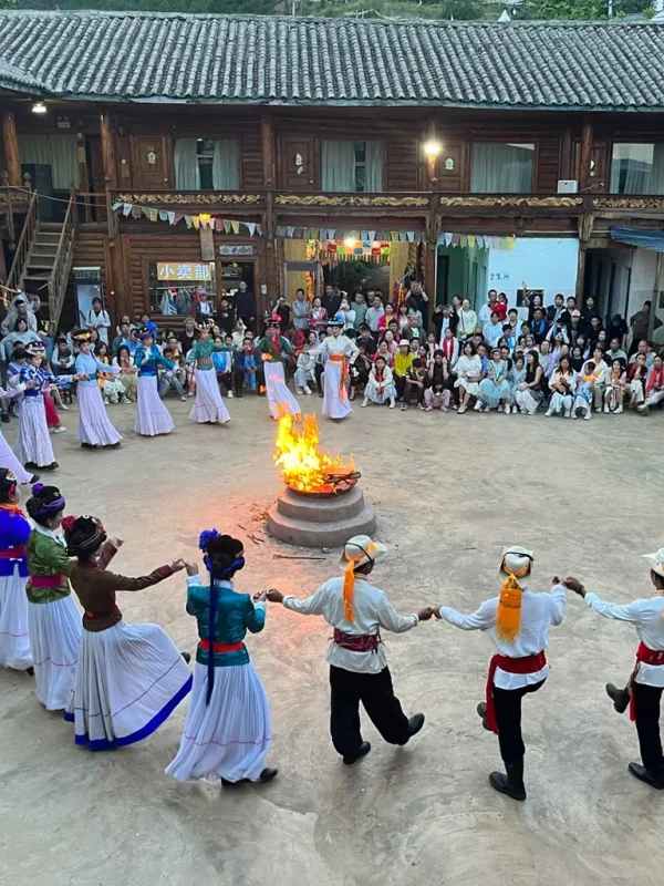 Mosuo Bonfire Party 瀘沽湖篝火晚會。身穿傳統摩梭服飾的男女手拉手圍成一圈,繞著巨大的點燃火坑起舞,身後是落座的觀眾和一座多層木建築。