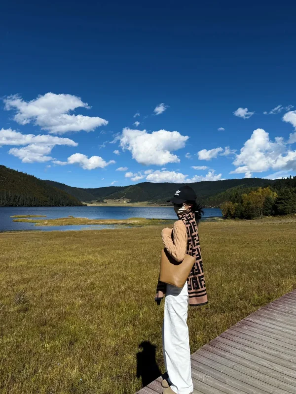 秋日的普達措屬都湖，在木棧道上側身回眸，金黃草甸、湖泊與藍天白雲相映，是香格里拉最溫柔的高原秋日畫面。