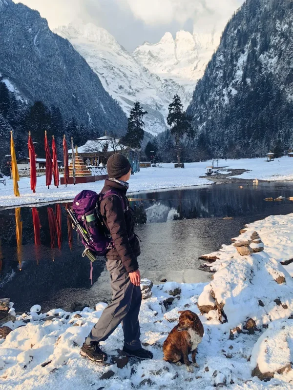 雨崩村冬日限定景觀，徒步者與狗狗站在結冰的湖畔，梅里雪山主峰倒映水中，宛如人間仙境。