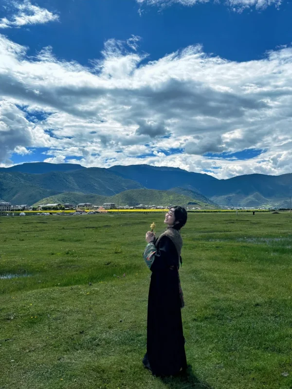 在納帕海草原上，穿著藏服、手持轉經輪，背後是遼闊的草甸、遠山與藍天白雲，是香格里拉最具代表性的旅行寫真畫面。