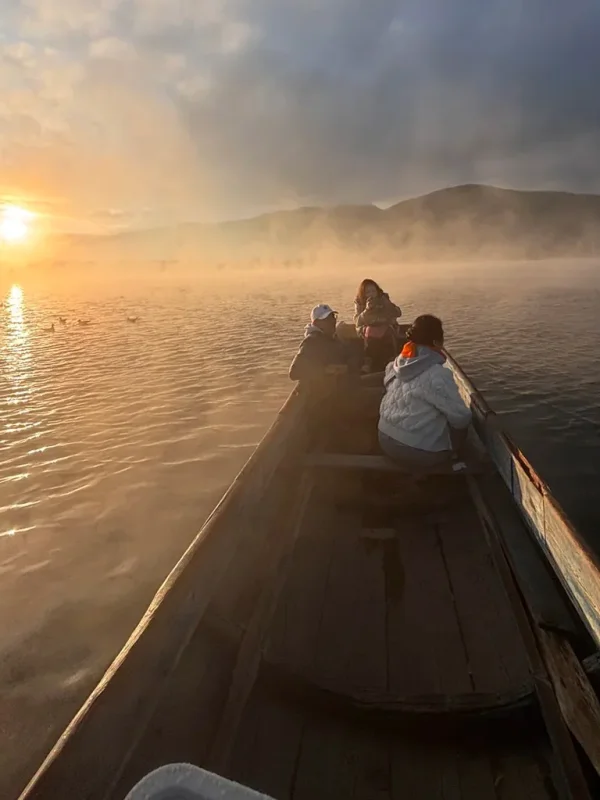 pig trough boats on Lugu Lake 2 雲南瀘沽湖的寧靜日出船遊。傳統木製豬槽船載著遊客,滑行在霧氣瀰漫的湖面,背景是金燦燦的日出與山景。